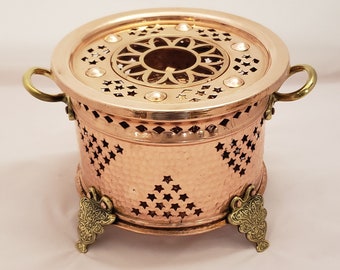 May include: A copper tea warmer with a decorative design and two handles. It has three ornate brass feet and a perforated top with a central opening.