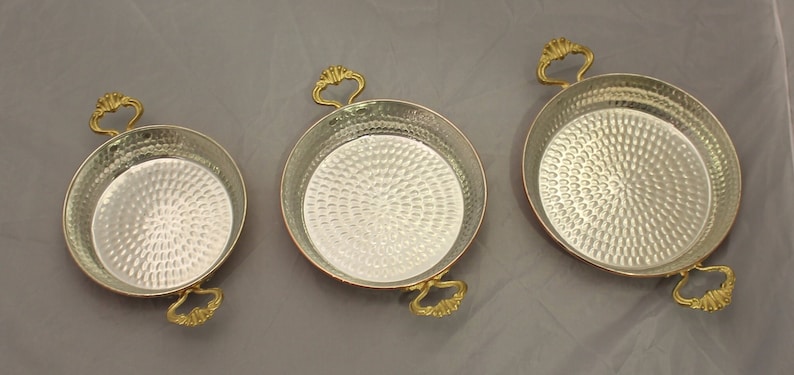 Op de afbeelding: Drie koperen serveerschalen met gehammerde oppervlakken en goudkleurige handvatten. De schalen zijn gerangschikt van klein naar groot.