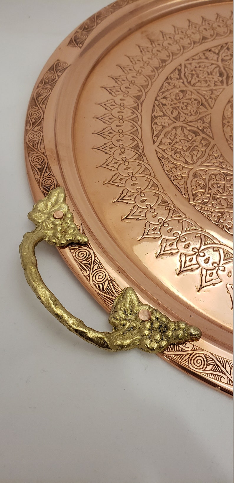 May include: A large, round copper serving tray with an intricate engraved design. The tray has two ornate gold handles with a grapevine motif.