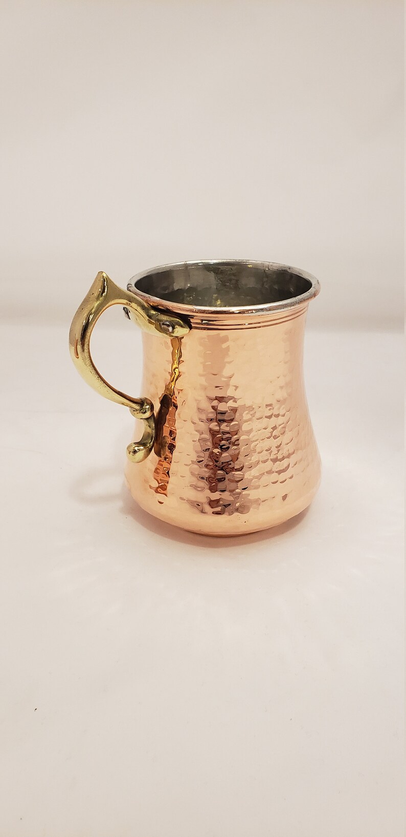 Pode incluir: Uma caneca de cobre martelada com uma al&ccedil;a de lat&atilde;o. A caneca tem um corpo arredondado e uma borda ligeiramente alargada.