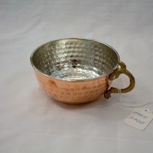 K&ouml;nnte beinhalten: Eine geh&auml;mmerte Kupferschale mit einem Messinggriff. Die Schale ist gl&auml;nzend und hat eine silberne Auskleidung. Die Schale ist mit "HAND MADE" beschriftet.
