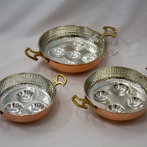 May include: Three copper-coloured escargot pans with hammered silver interiors and brass handles. Each pan has a round shape with six individual compartments for serving escargots. The pans are displayed on a white surface.