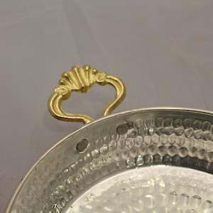 Op de afbeelding: Een gehamerde koperen serveerschaal met een gouden handvat. De schaal heeft een ronde vorm en een licht verhoogde rand.