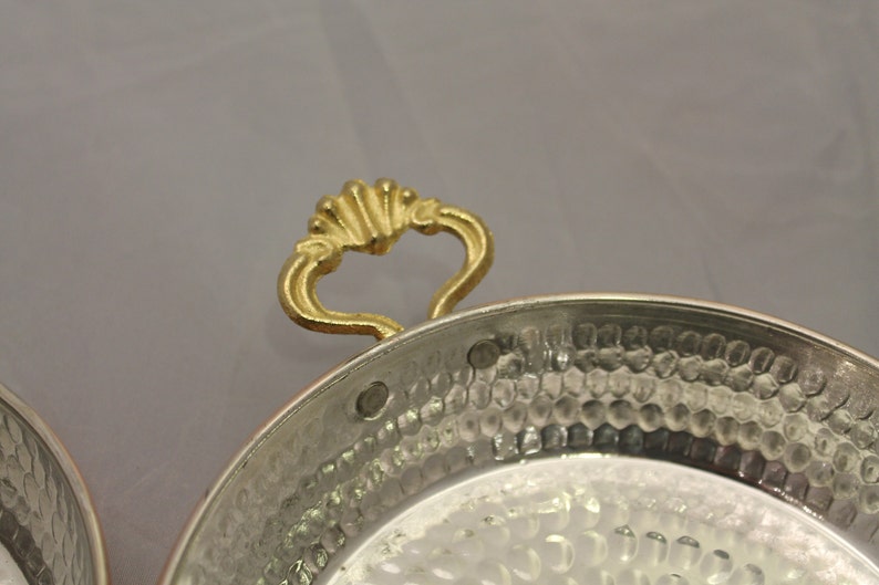 Op de afbeelding: Een gehamerde koperen serveerschaal met een gouden handvat. De schaal heeft een ronde vorm en een licht verhoogde rand.