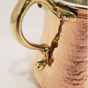 Pode incluir: Uma caneca de cobre martelada com uma al&ccedil;a de lat&atilde;o. A caneca tem um fundo arredondado e uma parte superior alargada. A al&ccedil;a &eacute; presa &agrave; caneca com dois rebites.
