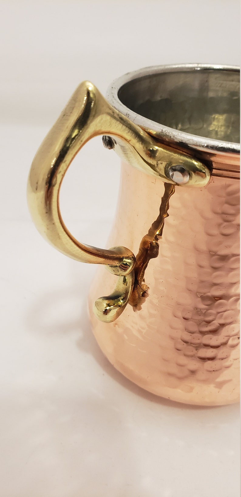Pode incluir: Uma caneca de cobre martelada com uma al&ccedil;a de lat&atilde;o. A caneca tem um fundo arredondado e uma parte superior alargada. A al&ccedil;a &eacute; presa &agrave; caneca com dois rebites.