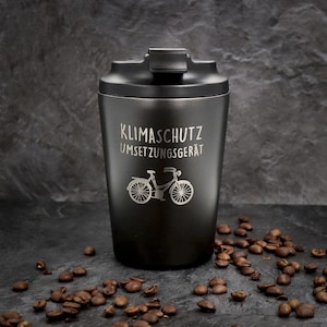Könnte beinhalten: Ein schwarzer Reisebecher mit Deckel, mit dem deutschen Text "KLIMASCHUTZ UMSETZUNGSGERÄT" und einer Fahrradillustration. Kaffeebohnen sind um den Becher verstreut. Der Becher ist für heiße oder kalte Getränke konzipiert.