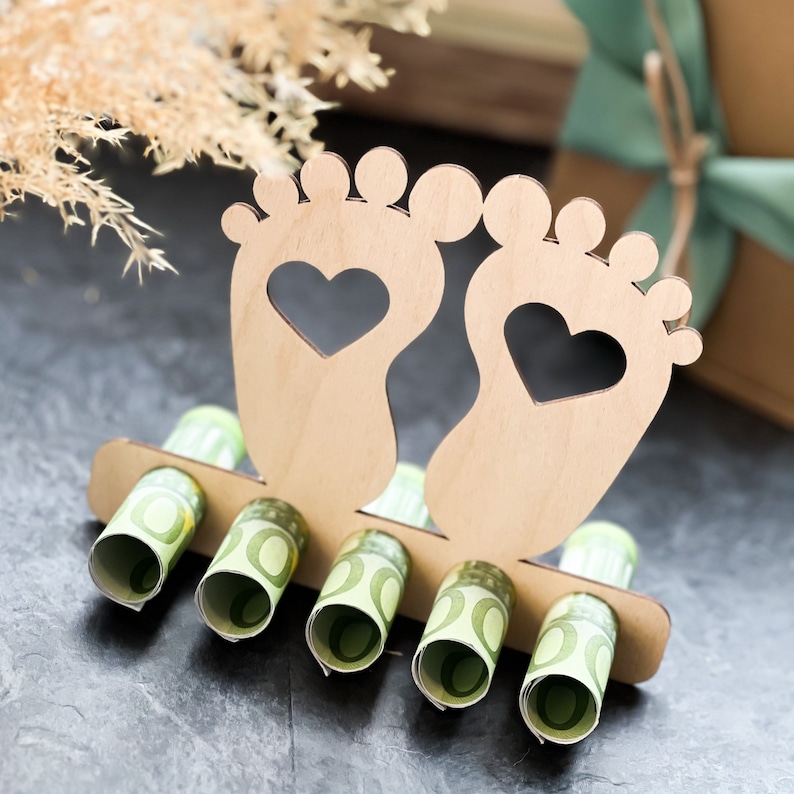 May include: A wooden baby feet-shaped money holder with heart cutouts. The holder has slots for rolled-up currency, with several rolls visible. The design is light brown, set against a dark background, suggesting a baby shower or new baby gift.