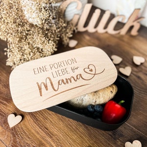 Peut inclure: Une boîte à lunch noire avec un couvercle en bois, partiellement ouverte pour révéler un petit pain, des myrtilles et un poivron rouge. Le couvercle est gravé des mots "EINE PORTION LIEBE für Mama" et d'un symbole de cœur.