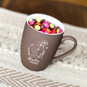 Könnte beinhalten: Eine braune Keramiktasse mit einem weißen Henkel und einem weißen Design eines Bären, der eine Blume hält. Die Tasse ist mit bunten Süßigkeiten gefüllt. Der Text "Munter Macher" ist in die Tasse graviert.