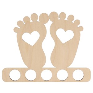 May include: A light-colored wooden baby feet-shaped decoration with a heart cut out of each foot. The decoration has a row of five circular holes at the bottom, likely for holding items. The design is simple and suitable for a nursery or baby-themed decor.