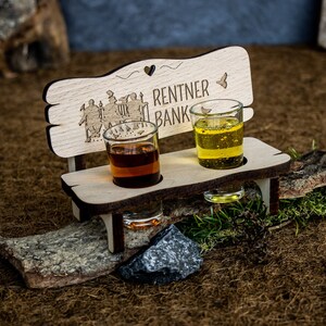 Könnte beinhalten: Ein Holz-Shotglashalter in Form einer Bank mit den Worten "RENTNER BANK" und einem Herz-Ausschnitt. Zwei Shotgläser sind mit bernsteinfarbenen und gelben Flüssigkeiten gefüllt. Die Bank ist mit geätzten Figuren und einem Schmetterling verziert.