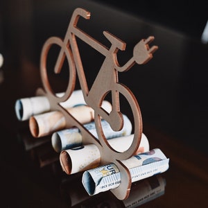 May include: Wooden cutout of an electric bicycle with a plug, holding rolled up euro bills.