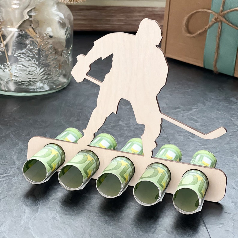 May include: Wooden hockey player money holder with five slots for rolled bills. The player is holding a hockey stick and is standing on a base with slots for bills.