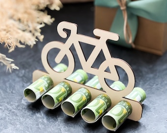 Könnte beinhalten: Ein hölzerner Fahrrad-Geldhalter mit sechs eingerollten Euro-Scheinen, die in die Schlitze gesteckt werden. Das Fahrrad ist in hellem Braun lackiert.
