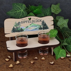 Könnte beinhalten: Holz-Schnapsglas-Halter in Form einer Bank mit den Worten "Gipfelglück" und einem Herz auf der Rückseite. Die Bank ist mit einem Aquarellbild einer Bergszene mit Bäumen und einem See verziert. Zwei Schnapsgläser sind mit einer rosa Flüssigkeit gefüllt.