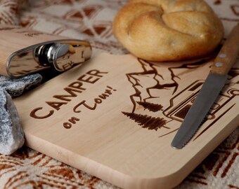 Könnte beinhalten: Ein Holzschneidebrett mit der Aufschrift "CAMPER on Tour!" und einer Berg- und Wohnwagen-Illustration. Eine Edelstahl-Flachmann und ein Brötchen sind ebenfalls vorhanden. Ein Messer liegt auf dem Brett.