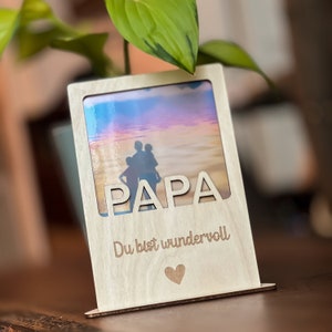 Könnte beinhalten: Ein Holz-Fotorahmen mit dem ausgeschnittenen Wort "PAPA" oben. Der Rahmen enthält ein Foto einer Familie am Strand. Unter dem Wort "PAPA" steht der Satz "Du bist wundervoll" und ein Herzsymbol.
