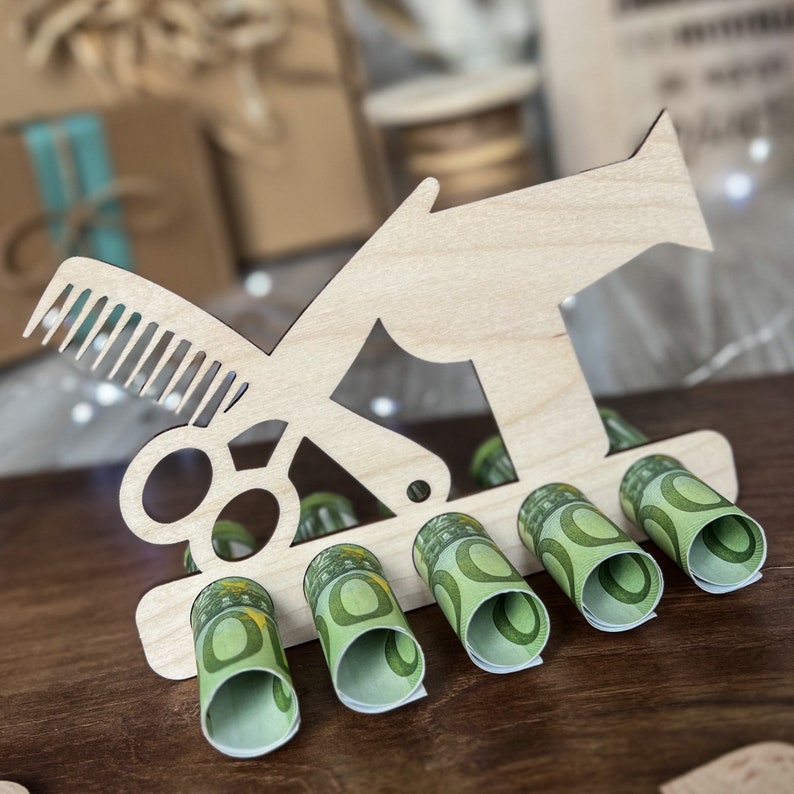 May include: A wooden barber-themed money holder with a comb and scissors design. The holder displays several rolled-up Euro banknotes. The wooden structure is light-colored, and the banknotes are green and white.