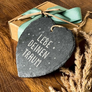 May include: A black heart-shaped slate sign with the text "LEBE DEINEN TRAUM" in white lettering. The sign is hanging from a brown twine string and is surrounded by a brown box with a green ribbon and dried grass.