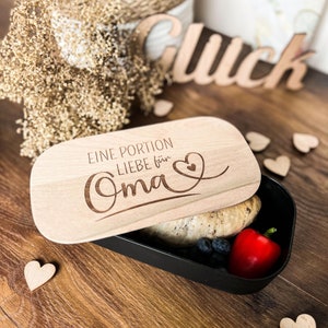Könnte beinhalten: Eine Holzlunchbox mit einem Deckel, auf dem "Eine Portion Liebe für Oma" mit einem Herz steht. Die Lunchbox ist gefüllt mit Essen, darunter Brot, Blaubeeren und eine rote Paprika.
