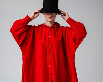 May include: A long, bright red hooded coat with gold-colored buttons and a matching drawstring. The coat has a relaxed fit and is paired with a black hat. The outfit is a bold fashion statement.