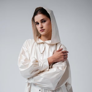 Puede incluir: Un impermeable con capucha de color crema con cierre de botones. El abrigo tiene mangas largas y una capucha con cordón. La modelo lleva el abrigo con los brazos cruzados. El fondo es blanco liso.