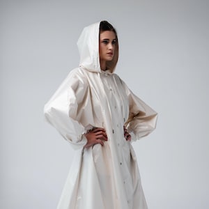 May include: A cream-colored hooded raincoat with a button-up front and long sleeves. The coat has a flared skirt and is worn over a white top. The model is wearing black shoes and white socks. The raincoat is a stylish outerwear garment.