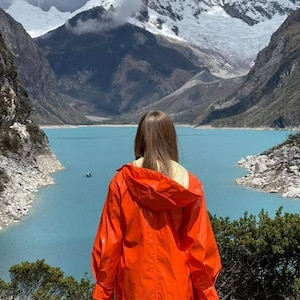 Puede incluir: Una persona con un impermeable con capucha de color naranja brillante se encuentra frente a un lago turquesa y montañas cubiertas de nieve. El paisaje incluye acantilados rocosos y vegetación verde. El cielo está nublado.