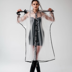 May include: A transparent raincoat with black trim and a hood. The raincoat is worn over a black dress and paired with black boots. The umbrella is held over the shoulders.