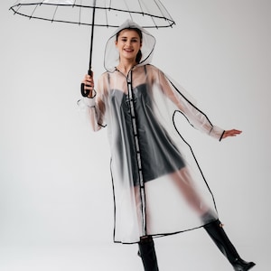 Puede incluir: Un impermeable transparente con ribetes negros y una capucha a juego. La persona sostiene un paraguas transparente con un mango negro. La persona lleva botas negras y un vestido negro. El fondo es blanco liso.