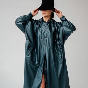 May include: A teal-colored raincoat with a hood and button-down front. The long coat features large pockets and is paired with a black hat. The outfit is a stylish, modern take on outerwear.