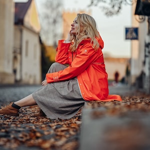 Puede incluir: Una mujer con un impermeable naranja brillante y una falda a cuadros se sienta en un bordillo con hojas caídas a su alrededor.