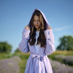 May include: A young person wearing a lavender hooded jacket with a belt. The jacket is unbuttoned and the hood is up. The person is standing in a field of lavender.