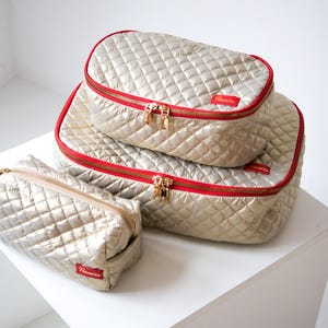 May include: Three quilted travel organizers in gold with red trim and gold zippers. The organizers are stacked on top of each other. The smallest organizer is on the bottom and the largest organizer is on top. The organizers have the word "Remonte" printed on them.