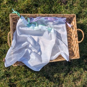 May include: A rectangular wicker basket with two handles contains several white mesh produce bags. The bags have green and lilac ribbon drawstrings. The basket rests on green grass.