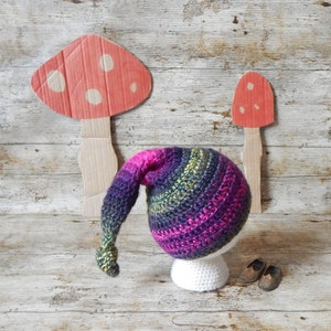May include: A purple, green and pink crocheted gnome hat with a long pointed tip. The hat is sitting on a white stand.