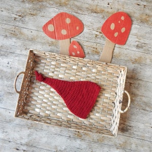 May include: A red crocheted hat sits in a woven basket. Three cardboard mushrooms with red caps and white spots are in the background. The basket is on a light wood-grain surface.