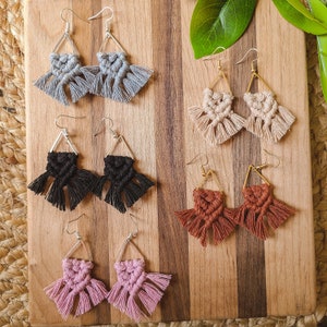 May include: Six pairs of handmade macrame earrings in various colors, including gray, black, beige, brown, and pink. Each pair features a triangular shape with fringe details.
