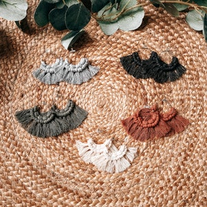 May include: Five pairs of handmade macrame earrings in various colors, including gray, black, brown, and white. The earrings are displayed on a woven surface.