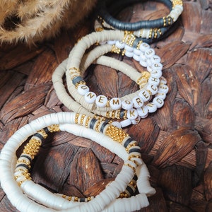 May include: Stack of handmade bracelets in white, black, and beige. Some bracelets have gold and black accents, and one spells out "ONE MORE KIND" with white letter beads. The bracelets are on a woven surface.