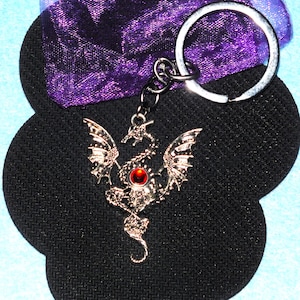 May include: Silver dragon keyring with a red gemstone. The dragon has detailed wings and a curled tail. The keyring is attached to a silver ring and hangs against a black, textured background.