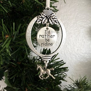 Puede incluir: Adorno navideño plateado con un encanto de caballo y una etiqueta redonda que dice "Prefiero estar montando".