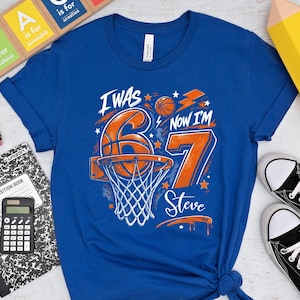 May include: A royal blue t-shirt with a basketball-themed graphic that reads "I WAS 6, NOW I'M 7 Steve". The image includes black sneakers, school supplies, and a wooden block sign that says "TEACH".