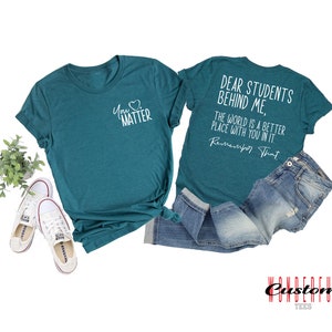 May include: Two teal blue t-shirts with white text. The front shirt says "You Matter" with a heart symbol. The back shirt says "Dear Students Behind Me, The World Is A Better Place With You In It. Remember That."