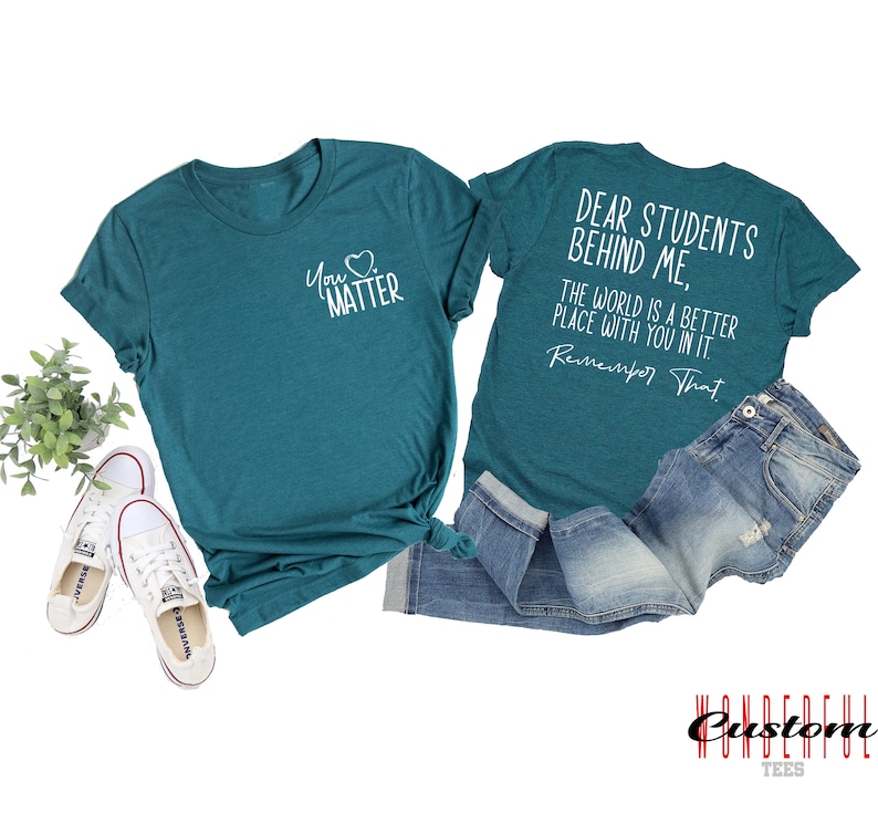 May include: Two teal blue t-shirts with white text. The front shirt says "You Matter" with a heart symbol. The back shirt says "Dear Students Behind Me, The World Is A Better Place With You In It. Remember That."