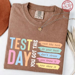 May include: A brown t-shirt with the words "TEST DAY YOU GOT THIS" in colorful block letters. Below the text are phrases like "rock the test" and "do your best". A silver necklace with a pendant is on the shirt.