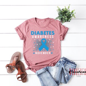 K&ouml;nnte beinhalten: Ein mauvefarbenes T-Shirt mit dem Text "Diabetes Awareness November" und einem blauen Band.