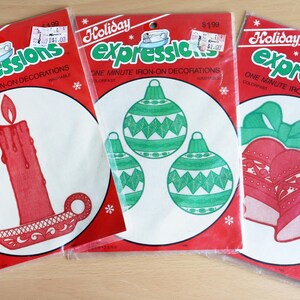 May include: Three packages of Holiday Expressions iron-on decorations. Each package is red with a white circle containing a different Christmas design: a red candle, green ornaments, and red bells with a green bow.