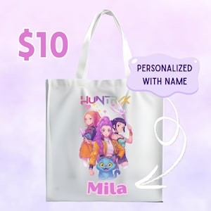 May include: White tote bag featuring a colorful illustration of three anime-style characters and a blue cat. The word "HUNTRAX" is printed above the characters, and the name "Mila" is below. The bag has white handles.